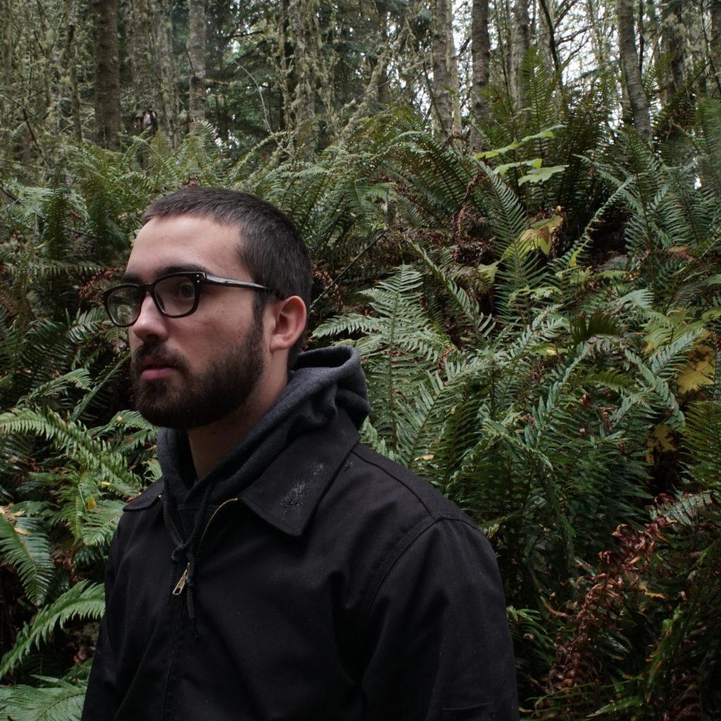 Indigenous man in a forest wearing a black jacket and black rimmed glasses.
