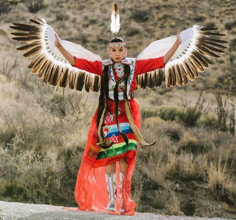 A Native American woman wearing eagle wings, a multi-colored dress and an eagle feather.