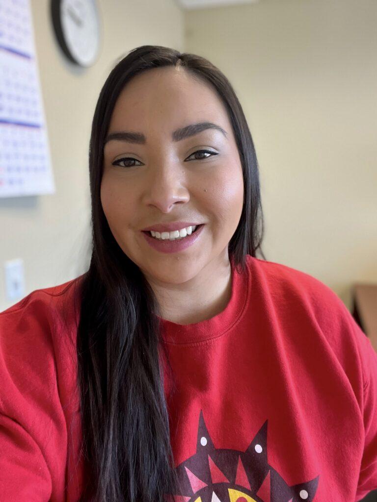 A close-up portrait of a long, brown- haired Native woman smiling. She is in an office setting, she is wearing a red shirt.