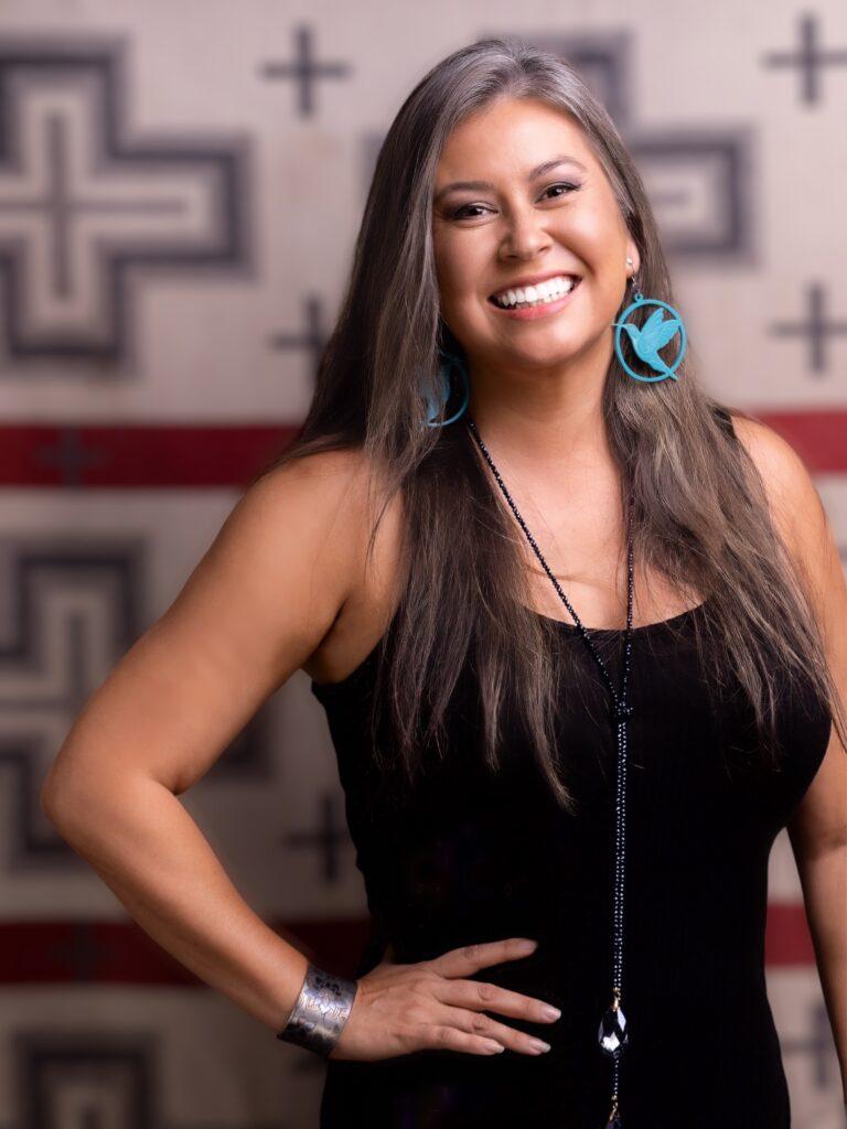 A smiling Indigenous woman with long hair and a black dress in front of a wall with plus and line designs