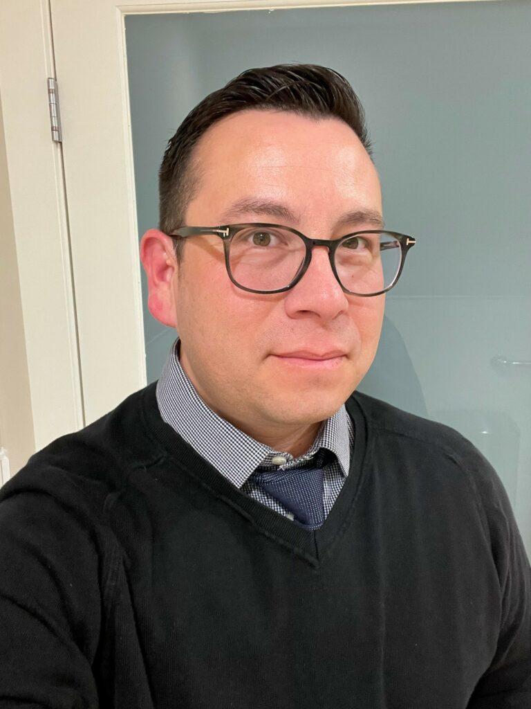 An Indigenous man with short hair and glasses wearing a sweater and collared shirt with a tie.