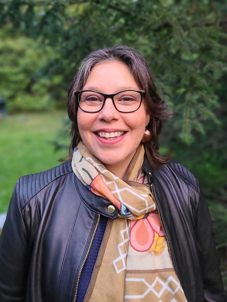 An Indigenous woman with glasses and leather jacket in front of a wooded area. She is smiling.