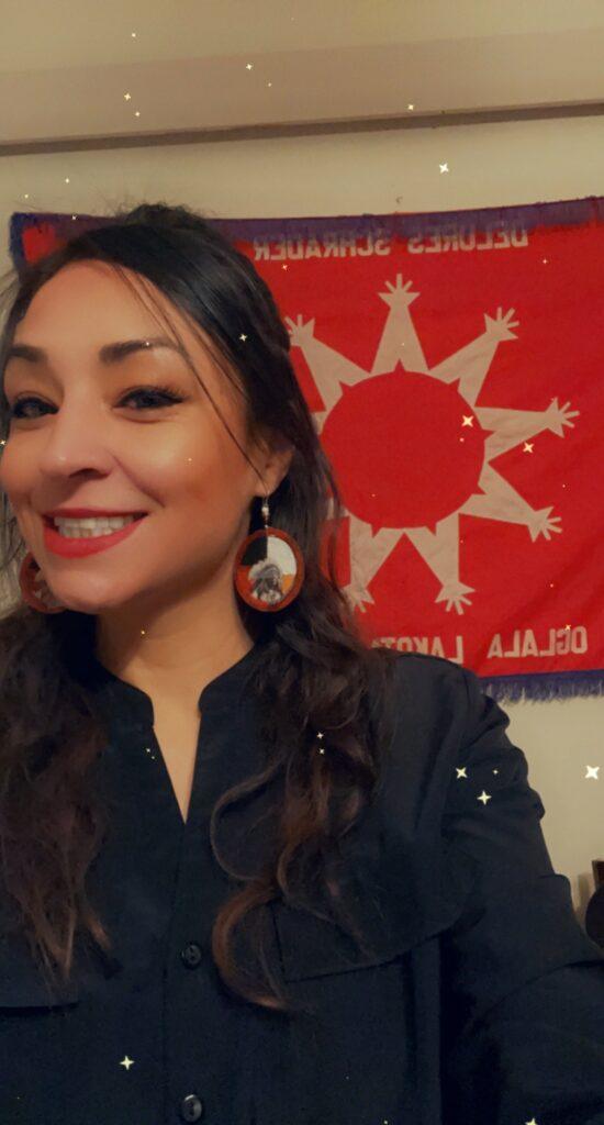 A smiling Native woman with long dark brown hair. She is wearing round beaded earrings and a navy blue shirt. There is a red tribal flag on the wall behind her.